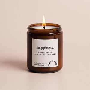 Può includere: Una candela accesa in un barattolo di vetro marrone con un'etichetta bianca. L'etichetta dice "happiness" ed elenca i profumi: bergamotto, lavanda, albero del tè al limone e arancia dolce. La candela è versata a mano con cera di soia e oli essenziali.