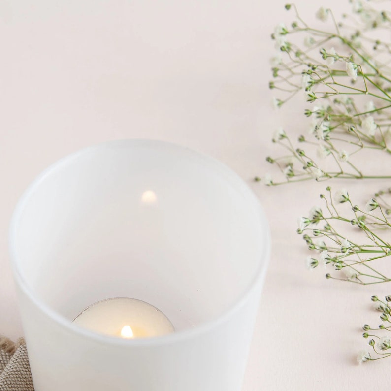 May include: A white frosted glass candle holder with a small white candle inside. The candle is lit and the flame is visible. The candle holder is on a light pink surface with a sprig of white baby's breath flowers.
