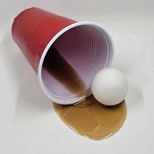 Op de afbeelding: Een rode plastic beker met een witte rand is omgekiept en morst een bruine vloeistof op een wit oppervlak. Een kleine witte bal ligt naast de mors. De beker heeft een geribbeld ontwerp.