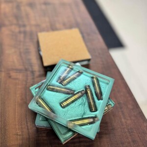 May include: Square resin coasters with a teal-green hue, embedded with brass bullet casings. The coasters are stacked on a wooden surface, with a brown square object in the background.