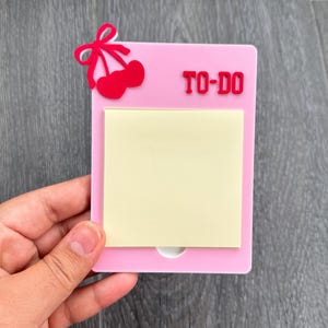May include: A pink acrylic To-Do list holder with a red cherry design and the words "TO-DO" in red. The holder has a yellow sticky note pad and a small white cutout at the bottom.
