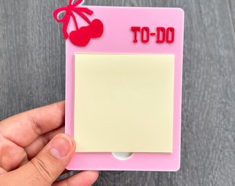 Pode incluir: Um suporte de lista de tarefas em acrílico rosa com um design de cereja vermelha e as palavras "TO-DO" em vermelho. O suporte tem um bloco de notas adesivas amarelo e um pequeno recorte branco na parte inferior.