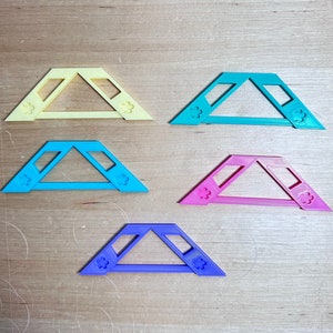 May include: Five colorful, geometric shapes in yellow, teal, blue, pink, and purple. Each shape has a triangular design with a flower-shaped cutout on each end. The shapes are arranged on a light brown wooden surface.