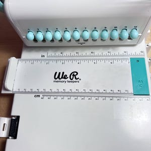 Pode incluir: Furador branco We R Memory Keepers Cinch com uma r&eacute;gua e um guia turquesa. O furador tem 12 furos numerados e uma marca central. A r&eacute;gua &eacute; marcada em polegadas e cent&iacute;metros.