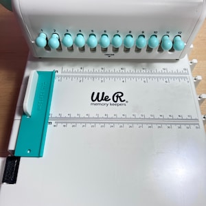 Pode incluir: M&aacute;quina de encaderna&ccedil;&atilde;o branca We R Memory Keepers Cinch com uma pega turquesa e uma r&eacute;gua com marcas at&eacute; 84 cm. A m&aacute;quina tem 12 furos de encaderna&ccedil;&atilde;o e uma marca central.