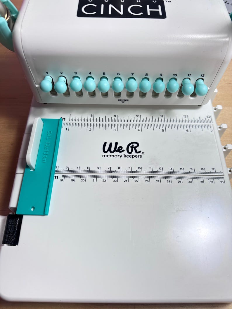 Pode incluir: M&aacute;quina de encaderna&ccedil;&atilde;o branca We R Memory Keepers Cinch com uma pega turquesa e uma r&eacute;gua com marcas at&eacute; 84 cm. A m&aacute;quina tem 12 furos de encaderna&ccedil;&atilde;o e uma marca central.