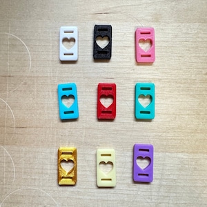 May include: Nine small, rectangular, heart-shaped charms in various colors: white, black, pink, blue, red, teal, gold, cream, and purple. Each charm has a heart cutout in the center and two small rectangular cutouts on the sides.