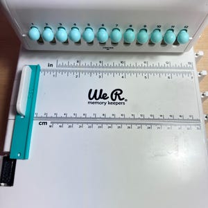 Pode incluir: Um cortador de papel branco We R Memory Keepers com uma r&eacute;gua azul e um deslizador turquesa. A r&eacute;gua tem marcas em polegadas e cent&iacute;metros. O cortador tem 12 ranhuras para perfurar furos.
