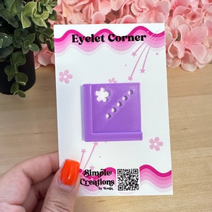 May include: Purple plastic eyelet corner tool with a flower and five holes. The tool is on a white card with the text "Eyelet Corner" and "Simple Creations by Kenja".