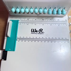 Pode incluir: Um cortador de papel branco We R Memory Keepers com uma r&eacute;gua azul e uma fileira de 12 pinos de perfura&ccedil;&atilde;o azuis. A r&eacute;gua &eacute; marcada em cent&iacute;metros e polegadas.