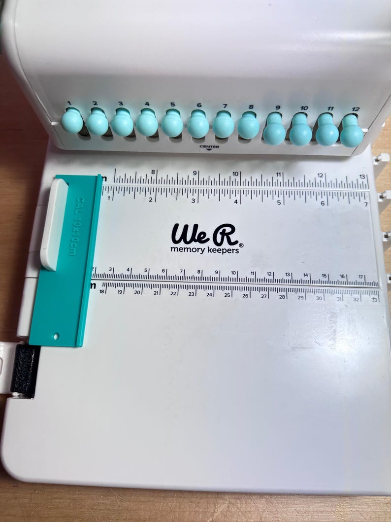 Pode incluir: Um cortador de papel branco We R Memory Keepers com uma r&eacute;gua azul e uma fileira de 12 pinos de perfura&ccedil;&atilde;o azuis. A r&eacute;gua &eacute; marcada em cent&iacute;metros e polegadas.