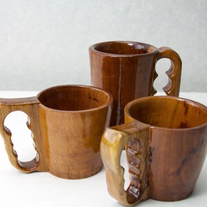 Op de afbeelding: Drie handgemaakte houten mokken met unieke handvatontwerpen. De mokken zijn gemaakt van een rijk, donkerbruin hout met een glanzende afwerking. De handvatten hebben een kenmerkend uitgesneden ontwerp, wat een vleugje artisticiteit toevoegt aan de rustieke esthetiek.