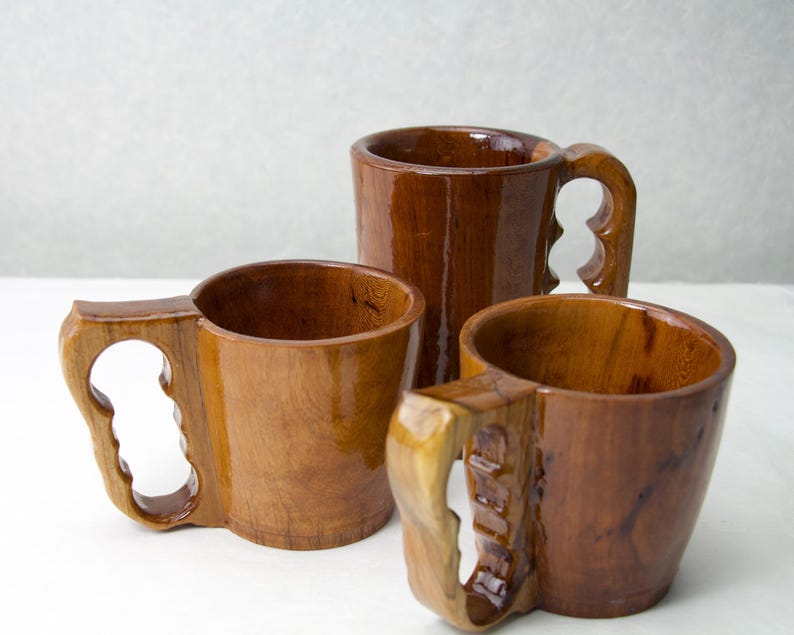 Op de afbeelding: Drie handgemaakte houten mokken met unieke handvatontwerpen. De mokken zijn gemaakt van een rijk, donkerbruin hout met een glanzende afwerking. De handvatten hebben een kenmerkend uitgesneden ontwerp, wat een vleugje artisticiteit toevoegt aan de rustieke esthetiek.