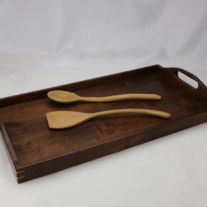 May include: A rectangular wooden serving tray with two wooden spoons. The tray is made of dark wood with a light wood border. The spoons are made of light wood and have a natural finish.