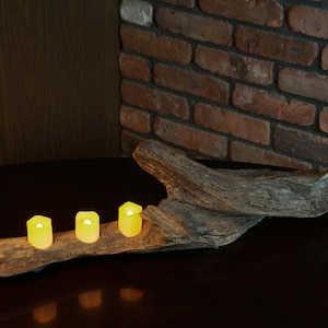 Puede incluir: Un portavelas rústico de madera de deriva con tres velas de té encendidas. El portavelas está sobre una mesa marrón oscura con una pared de ladrillo en el fondo.