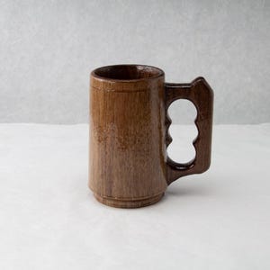 Op de afbeelding: Een bruine houten bierpul met een uniek handvat. De mok heeft een cilindrisch lichaam en een stevige basis. Het handvat heeft drie vingergaten voor een comfortabele grip. De pul is gemaakt van hout en heeft een natuurlijke afwerking.