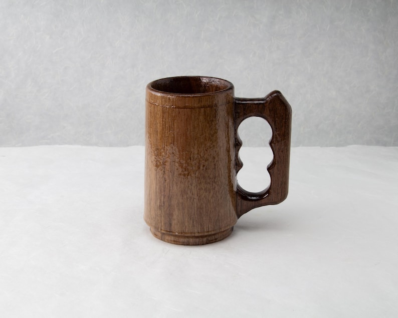 Op de afbeelding: Een bruine houten bierpul met een uniek handvat. De mok heeft een cilindrisch lichaam en een stevige basis. Het handvat heeft drie vingergaten voor een comfortabele grip. De pul is gemaakt van hout en heeft een natuurlijke afwerking.