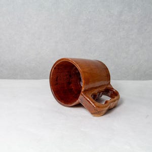Op de afbeelding: Een handgemaakte bruine keramische mok met een uniek handvatontwerp. De mok heeft een glanzende afwerking en een rijke, aardse tint. De binnenkant van de mok is een donkerdere bruintint, wat een contrast cre&euml;ert. Geschikt voor warme of koude dranken.