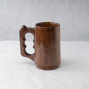 Op de afbeelding: Een handgemaakte houten mok met een uniek handvatontwerp. De mok is gemaakt van donkerbruin hout met een glanzende afwerking. Het handvat heeft drie cirkelvormige uitsparingen, wat een onderscheidend tintje geeft. De mok is geschikt voor dranken.
