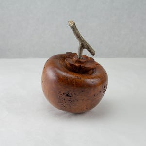 May include: A wooden sculpture of a persimmon fruit with a small twig stem. The sculpture is carved with intricate details and has a rich brown color.