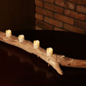 Puede incluir: Un portavelas de madera rústico con cuatro velas encendidas. El portavelas está hecho de una rama natural de madera flotante y tiene un color marrón natural.