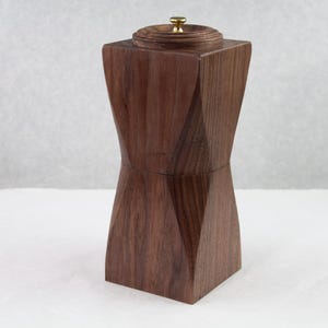 Könnte beinhalten: Ein handgefertigter Pfeffermühle aus Holz mit geometrischem Design. Das dunkelbraune Holz wird durch einen kleinen goldfarbenen Knopf auf der Oberseite akzentuiert. Die Mühle hat eine quadratische Basis und einen breiteren Mittelteil.