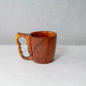 Op de afbeelding: Een handgemaakte houten mok met een uniek handvatontwerp. De mok is gemaakt van donkerbruin hout met een lichter gekleurd handvat met drie vingergaten. De binnenkant van de mok is een rijk, warm bruin.