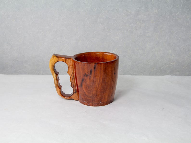 Op de afbeelding: Een handgemaakte houten mok met een uniek handvatontwerp. De mok is gemaakt van donkerbruin hout met een lichter gekleurd handvat met drie vingergaten. De binnenkant van de mok is een rijk, warm bruin.