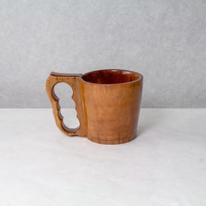 Op de afbeelding: Een handgemaakte houten mok met een uniek handvatontwerp. De mok is gemaakt van lichtbruin hout en heeft een uitgesneden handvat met drie vingergaten. De binnenkant van de mok is een donkerdere bruintint. De mok staat op een wit oppervlak.