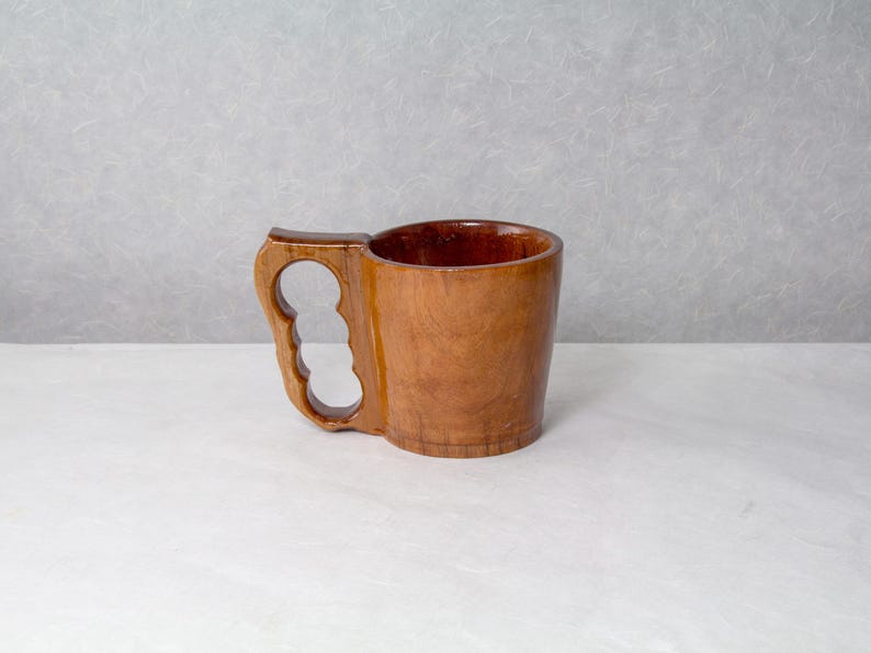 Op de afbeelding: Een handgemaakte houten mok met een uniek handvatontwerp. De mok is gemaakt van lichtbruin hout en heeft een uitgesneden handvat met drie vingergaten. De binnenkant van de mok is een donkerdere bruintint. De mok staat op een wit oppervlak.