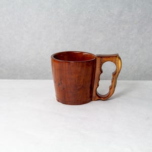 Op de afbeelding: Een handgemaakte houten mok met een uniek handvatontwerp. De mok is gemaakt van donkerbruin hout met een gepolijste afwerking, die de natuurlijke nerf benadrukt. Het handvat heeft een opvallende uitsparing, wat bijdraagt aan de rustieke charme. Hoogte ca. 10 cm.