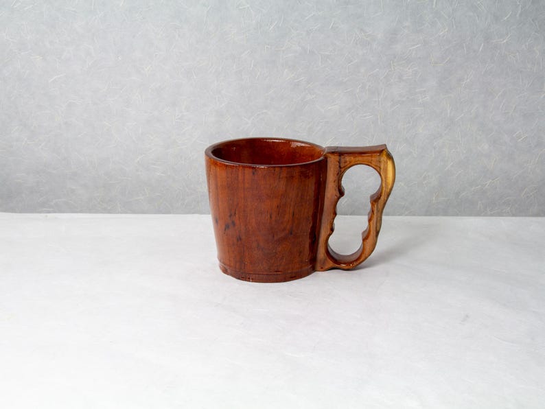 Op de afbeelding: Een handgemaakte houten mok met een uniek handvatontwerp. De mok is gemaakt van donkerbruin hout met een gepolijste afwerking, die de natuurlijke nerf benadrukt. Het handvat heeft een opvallende uitsparing, wat bijdraagt aan de rustieke charme. Hoogte ca. 10 cm.