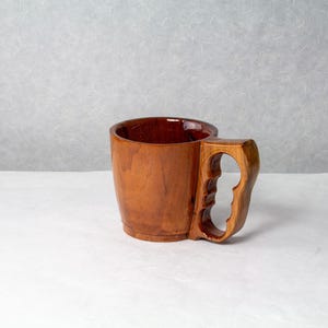 Op de afbeelding: Een handgemaakte houten mok met een uniek handvatontwerp. De mok is gemaakt van gepolijst hout met een rijke, warme bruine kleur. Het handvat is uitgesneden met vingergroeven voor een comfortabele grip. De binnenkant van de mok heeft een donkerdere, roodbruine tint.