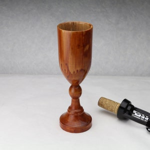 May include: A handcrafted wooden goblet with a stem and a wide bowl. The goblet is a warm brown color with visible wood grain. A corkscrew with a cork is next to the goblet.