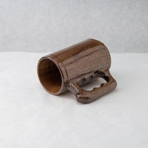 Op de afbeelding: Een handgemaakte houten mok met een donkerbruine afwerking. De mok heeft een cilindrisch lichaam en een stevige handgreep met drie vingeruitsparingen. De binnenkant van de mok is lichter bruin, in contrast met de buitenkant. De mok staat tegen een neutrale achtergrond.