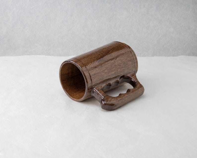 Op de afbeelding: Een handgemaakte houten mok met een donkerbruine afwerking. De mok heeft een cilindrisch lichaam en een stevige handgreep met drie vingeruitsparingen. De binnenkant van de mok is lichter bruin, in contrast met de buitenkant. De mok staat tegen een neutrale achtergrond.