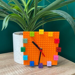 Może przedstawiać: Pomarańczowy zegar Lego z kolorowymi klockami Lego wok&oacute;ł tarczy. Zegar ma czarne wskaz&oacute;wki i czerwoną wskaz&oacute;wkę sekundową.