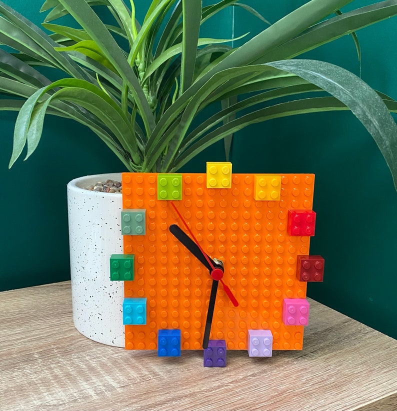 Może przedstawiać: Pomarańczowy zegar Lego z kolorowymi klockami Lego wok&oacute;ł tarczy. Zegar ma czarne wskaz&oacute;wki i czerwoną wskaz&oacute;wkę sekundową.