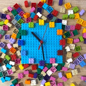 Może przedstawiać: Niebieska tarcza zegara Lego z czarnymi wskaz&oacute;wkami, otoczona kolorowymi klockami Lego. Tarcza zegara jest wykonana z małych niebieskich kwadrat&oacute;w.