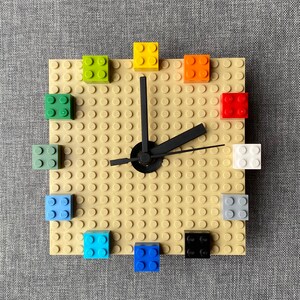 Może przedstawiać: Zegar wykonany z klock&oacute;w LEGO z czarną wskaz&oacute;wką i beżową podstawą. Tarcza zegara jest wykonana z małych beżowych klock&oacute;w, a cyfry są reprezentowane przez klocki LEGO w r&oacute;żnych kolorach.