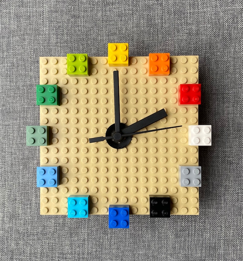 Może przedstawiać: Zegar wykonany z klock&oacute;w LEGO z czarną wskaz&oacute;wką i beżową podstawą. Tarcza zegara jest wykonana z małych beżowych klock&oacute;w, a cyfry są reprezentowane przez klocki LEGO w r&oacute;żnych kolorach.