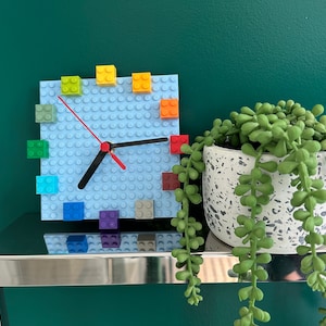 Może przedstawiać: Niebieski zegar Lego z kolorowymi klockami Lego wok&oacute;ł tarczy. Zegar znajduje się na srebrnej p&oacute;łce z zieloną ścianą w tle. Roślina doniczkowa z sukulentami znajduje się na pierwszym planie.