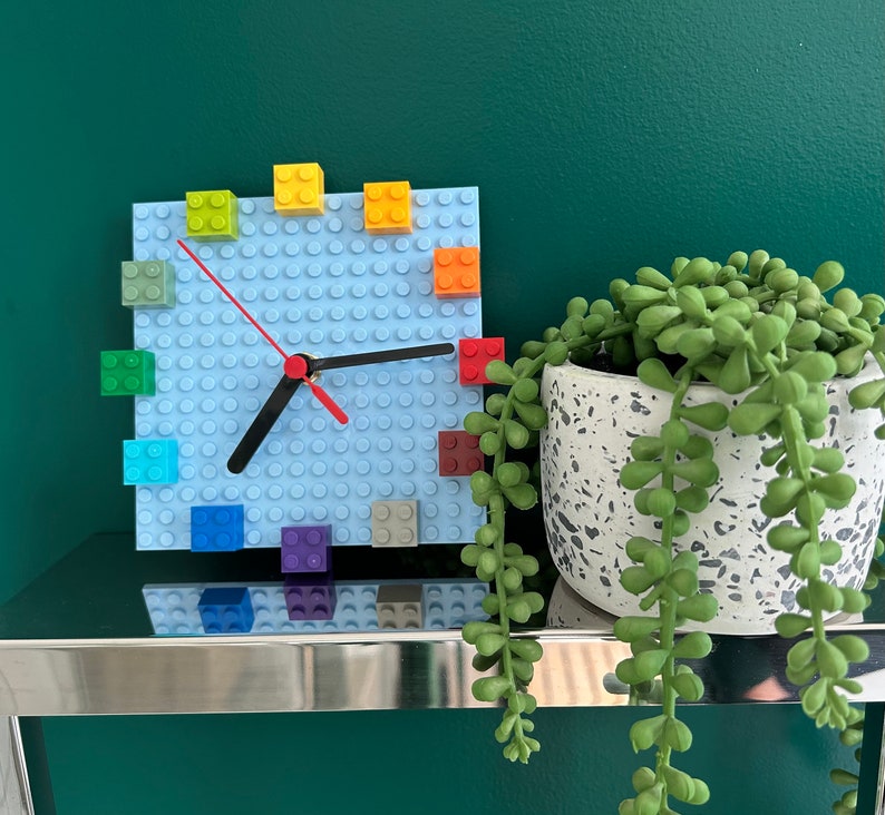 Może przedstawiać: Niebieski zegar Lego z kolorowymi klockami Lego wok&oacute;ł tarczy. Zegar znajduje się na srebrnej p&oacute;łce z zieloną ścianą w tle. Roślina doniczkowa z sukulentami znajduje się na pierwszym planie.
