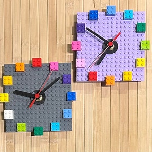 Może przedstawiać: Dwa zegary Lego, jeden z podstawą fioletową, a drugi z podstawą szarą. Oba zegary mają kolorowe klocki Lego wok&oacute;ł krawędzi i czarną wskaz&oacute;wkę zegara.