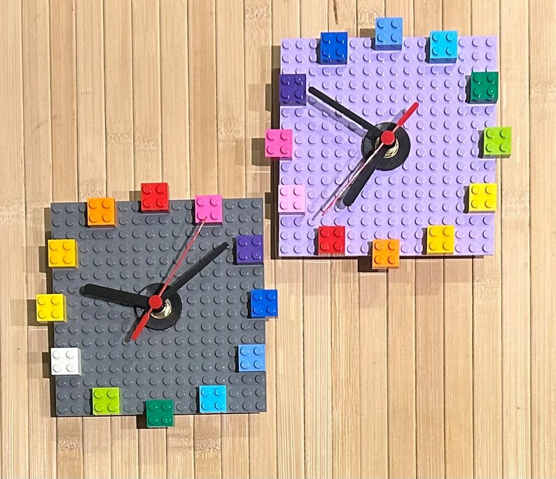 Może przedstawiać: Dwa zegary Lego, jeden z podstawą fioletową, a drugi z podstawą szarą. Oba zegary mają kolorowe klocki Lego wok&oacute;ł krawędzi i czarną wskaz&oacute;wkę zegara.