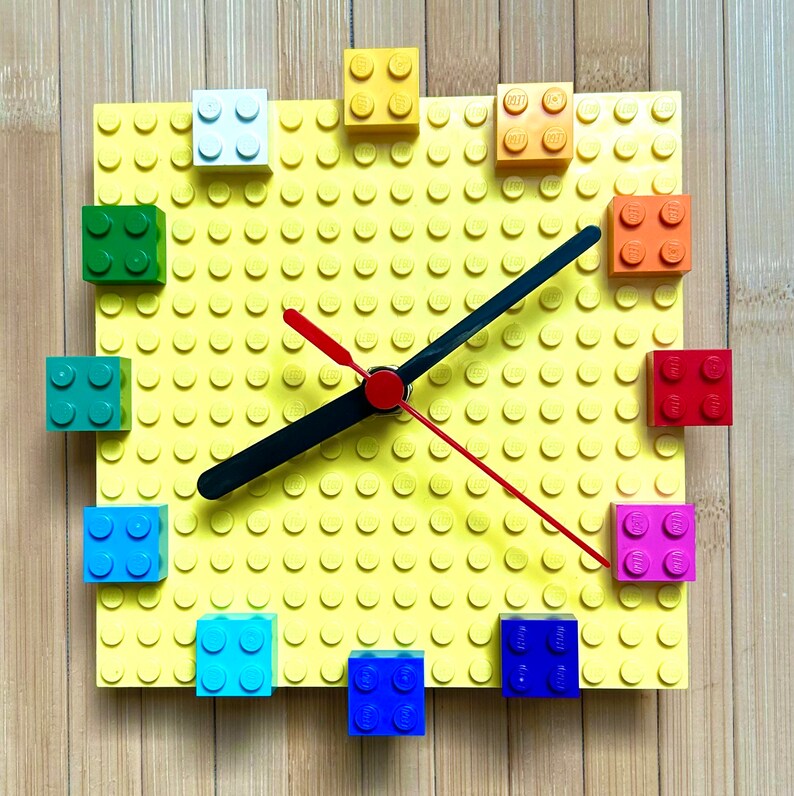 Może przedstawiać: Ż&oacute;łty zegar Lego z czarną i czerwoną wskaz&oacute;wką. Tarcza zegara jest wykonana z klock&oacute;w Lego i ma r&oacute;żne kolorowe klocki wok&oacute;ł krawędzi.