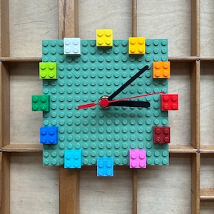 Może przedstawiać: Zielony zegar Lego z czarną wskaz&oacute;wką minutową i czerwoną wskaz&oacute;wką godzinową. Tarcza zegara jest wykonana z klock&oacute;w Lego, a cyfry są reprezentowane przez kolorowe klocki Lego.