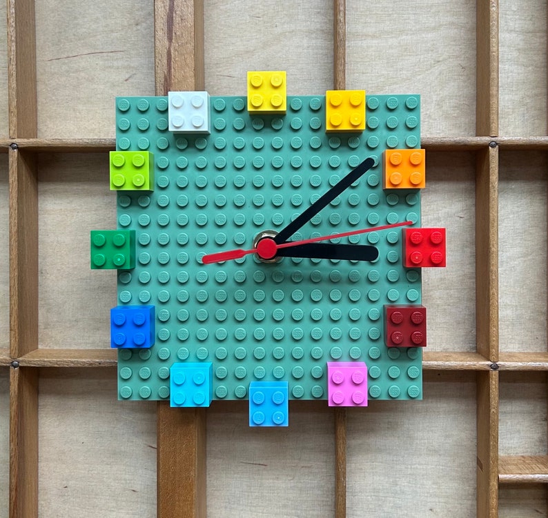 Może przedstawiać: Zielony zegar Lego z czarną wskaz&oacute;wką minutową i czerwoną wskaz&oacute;wką godzinową. Tarcza zegara jest wykonana z klock&oacute;w Lego, a cyfry są reprezentowane przez kolorowe klocki Lego.