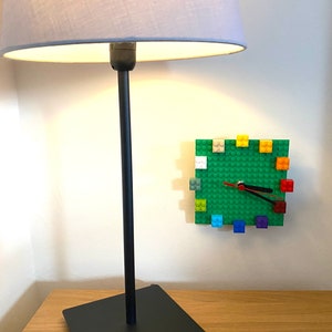 Może przedstawiać: Czarna lampa z szarym kloszem stoi na drewnianym stole. Zielony zegar Lego z kolorowymi klockami Lego jest zamontowany na ścianie za lampą.
