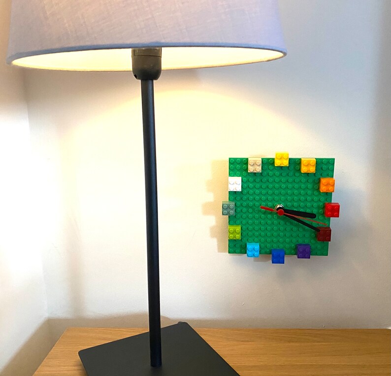 Może przedstawiać: Czarna lampa z szarym kloszem stoi na drewnianym stole. Zielony zegar Lego z kolorowymi klockami Lego jest zamontowany na ścianie za lampą.